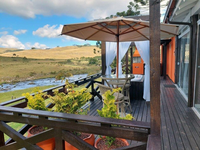 Deck de madeira com vista para uma paisagem deslumbrante com um rio. Uma mesa com guarda-sol, cadeiras e vasos de plantas estão dispostos no deck.