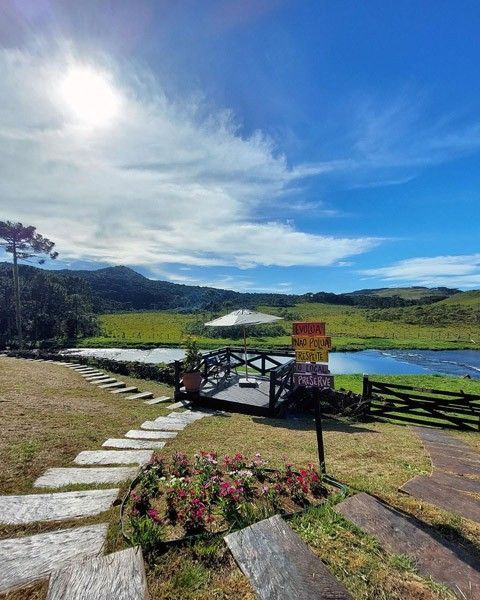 Paisagem pitoresca com um caminho de pedra, ponte de madeira, flores e um rio sob um céu azul brilhante.