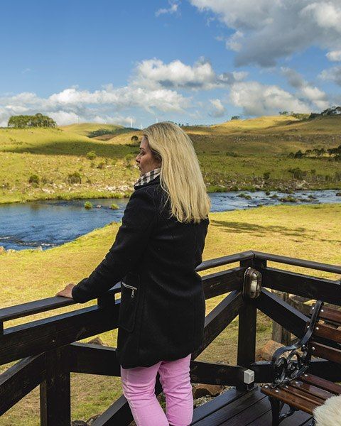 Mulher em pé num deck de madeira, olhando para um rio e colinas gramadas sob um céu azul.