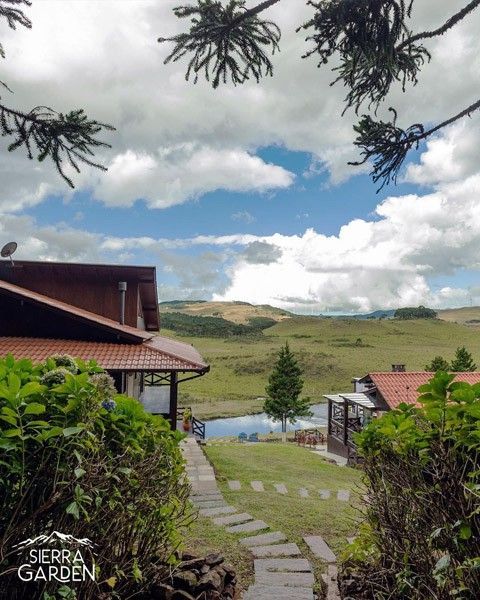 Um caminho de pedra leva a casas com telhados alaranjados, com vista para um campo gramado e um lago, sob um céu nublado.