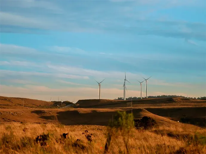 Turbinas eólicas em uma crista numa paisagem seca e gramada sob um céu azul.