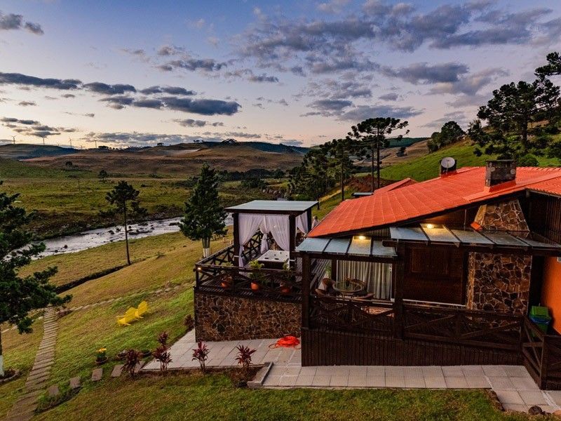 Cabana pitoresca com telhado laranja e fachada de pedra, com vista para um rio e colinas sob um céu nublado.