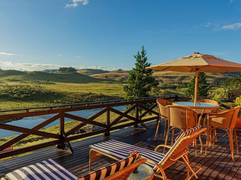 Deck de madeira com cadeiras, mesa e guarda-sol com vista para uma paisagem verdejante sob um céu azul.