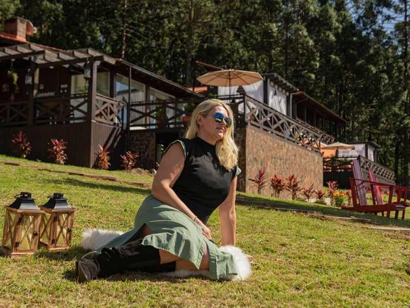 Mulher de óculos escuros sentada na grama, perto de uma cabana em um local ensolarado.