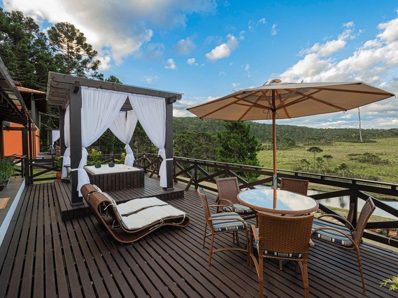 Deck de madeira com espreguiçadeira, mesa com cadeiras e gazebo com cortinas, com vista para um prado.