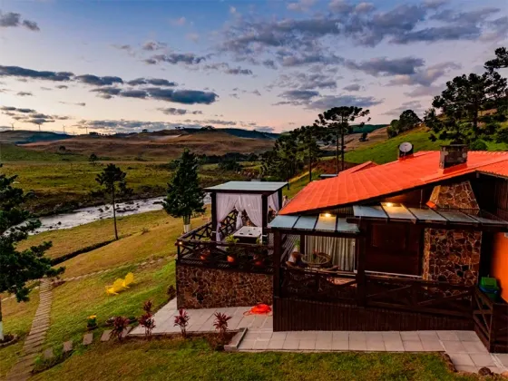 Cabana com telhado laranja, fachada de pedra e deck com vista para uma paisagem rural ao pôr do sol.