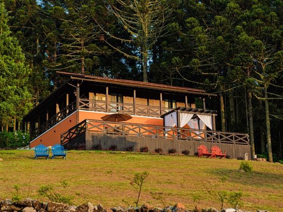 Cabana com exterior laranja e corrimão de madeira, rodeada por árvores. Cadeiras azuis e vermelhas estão no gramado.