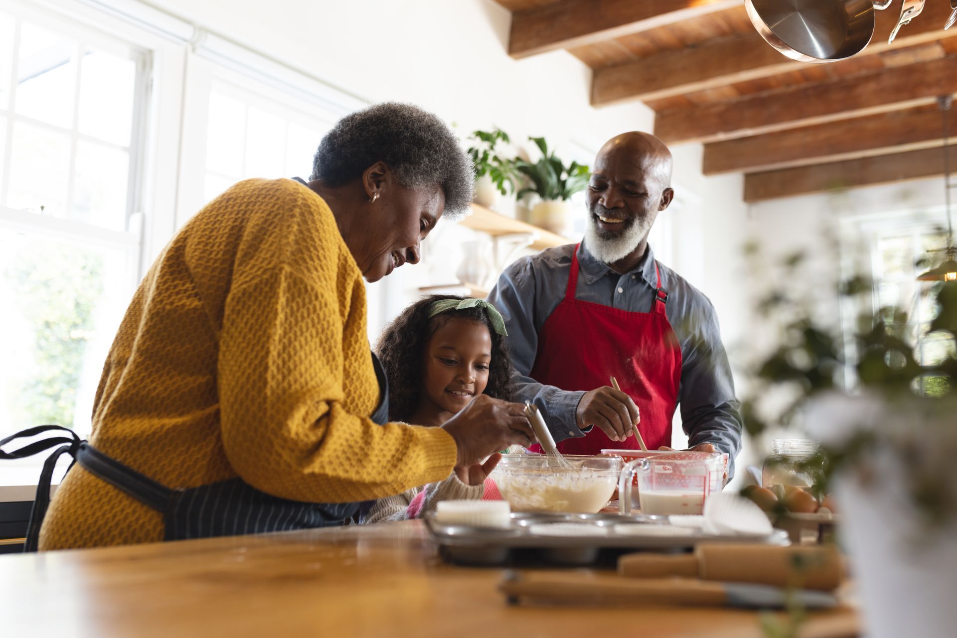 Two adults and a child wearing aprons mix ingredients in bowls while standing together in a sunny kitchen.