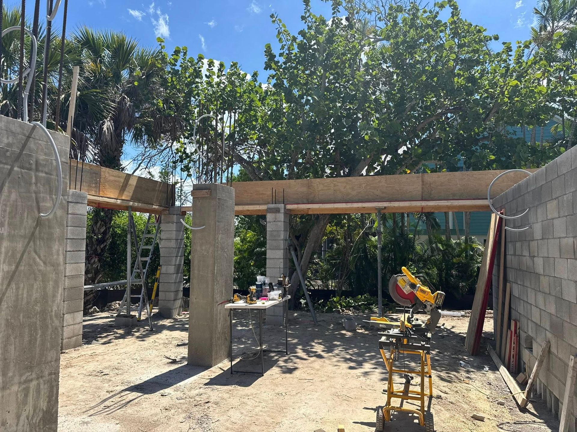 Outdoor patio under construction with stone walls, wooden beams, and chairs in a sunny yard