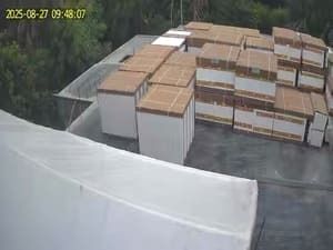Outdoor industrial yard with stacks of boxed pallets on wet pavement and a timestamp in the corner