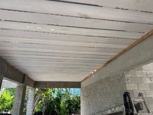 Covered patio ceiling with white beams and exposed gray walls, opening to greenery outside