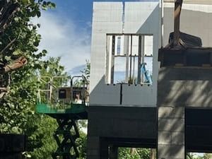 Construction lift beside a gray building, with a worker platform elevated near trees and blue sky.