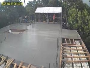 Rooftop construction site with a wet concrete slab, exposed framing, and workers under a small roofed structure.