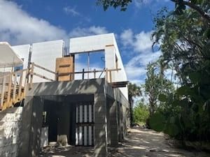 Partially built house with white walls and a wooden balcony beside a dirt path and trees