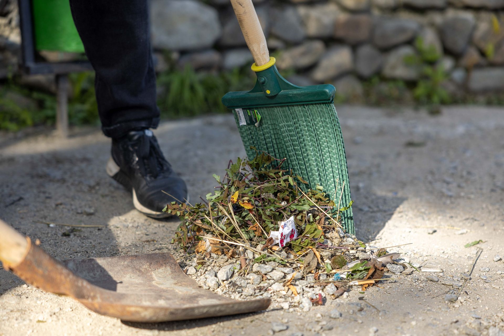 Person using a green litter picker to collect trash from gravel near a stone wall.