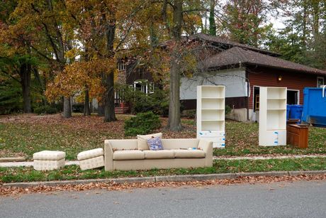 Discarded furniture on a curb in front of a house, with autumn trees and a blue dumpster nearby.