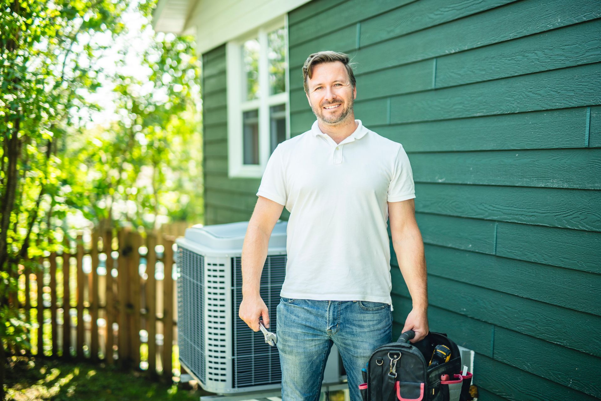 A technician holding a toolkit working on an air conditioning unit outdoors.