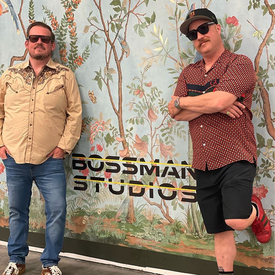 Two men posing in front of a Bossman Studios mural, both wearing sunglasses and casual clothes.