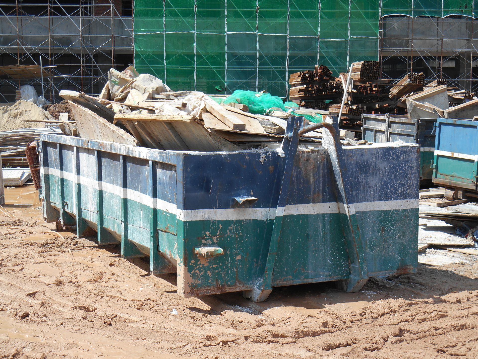 Large blue-green dumpster filled with construction debris on a muddy construction site