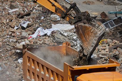 Excavator dumping rubble into an orange dumpster at a demolition site