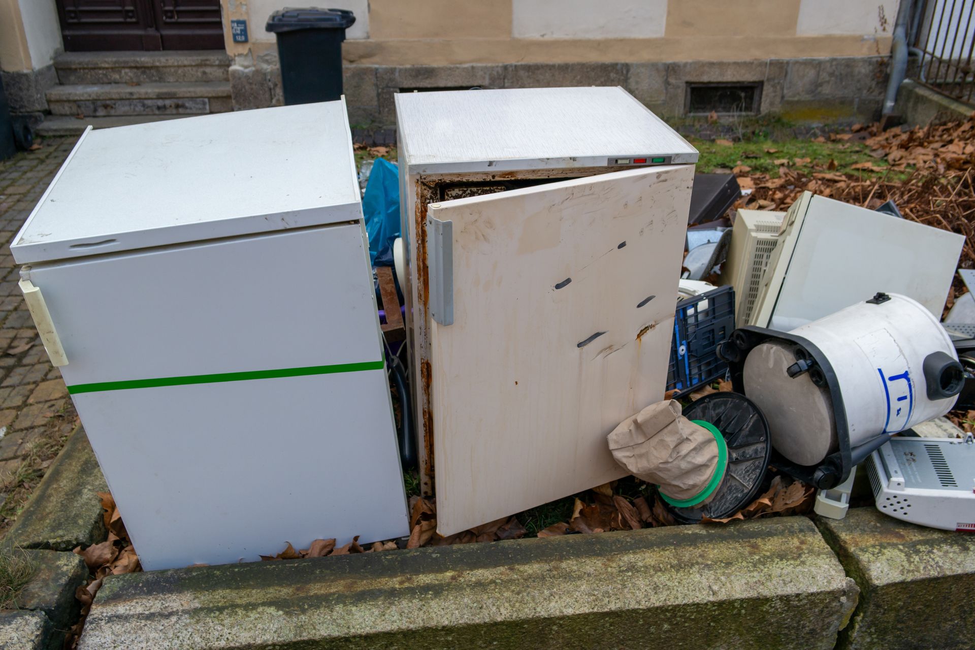Piles of discarded furniture and appliances on a curb, including white cabinets and a broken fan.