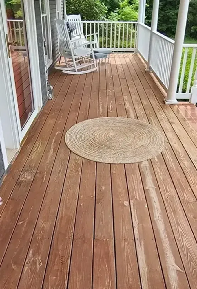 Wooden porch with white railings, gray siding, potted flowers, and a round woven rug.