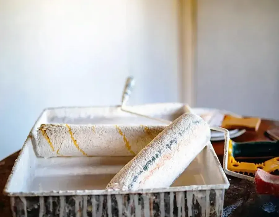 Paint roller tray with white paint and a roller on a table, showing yellow paint drips and a paint can nearby