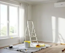 Bright empty room during painting, with ladder, drop cloths, and paint supplies by a window.