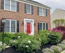 Two-story brick house with red front door and white trim, framed by lush green shrubs and pink flowers.