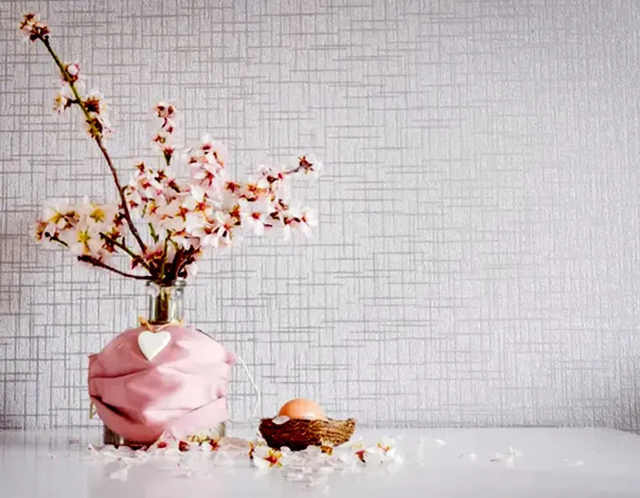 Pink vase with flowering branches and a gold teapot on a white table against a textured gray wall
