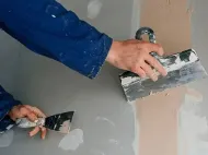 Person applying gray plaster to a wall with two trowels.