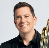 Smiling man in a black shirt beside a trombone.