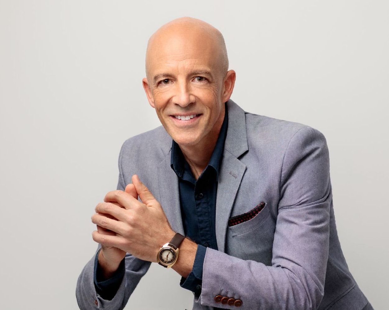 A bald man wearing a suit and a watch is sitting with his hands folded.