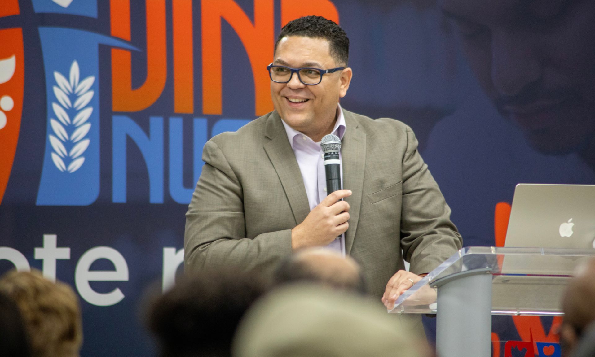 Dr. Peter Rios smiling, holding a microphone, speaking at a podium with a backdrop.