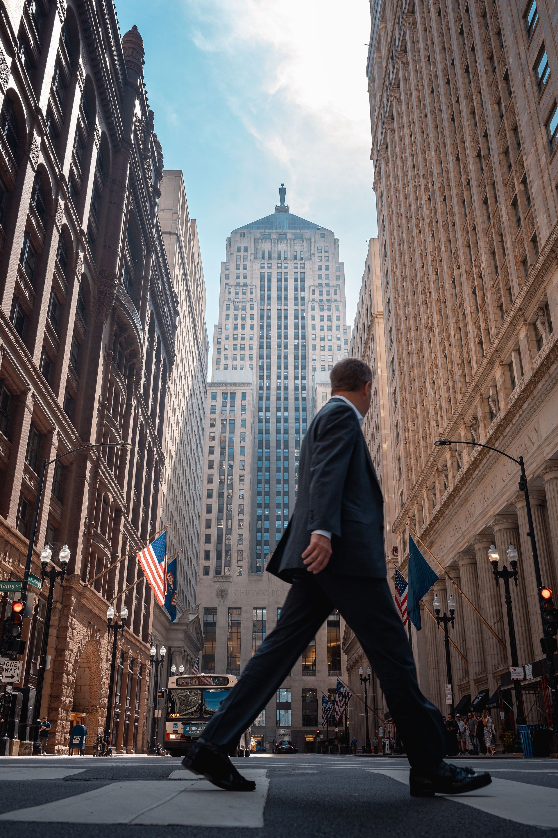 Man in a suit walking across a city street between tall buildings, with a skyscraper in the background