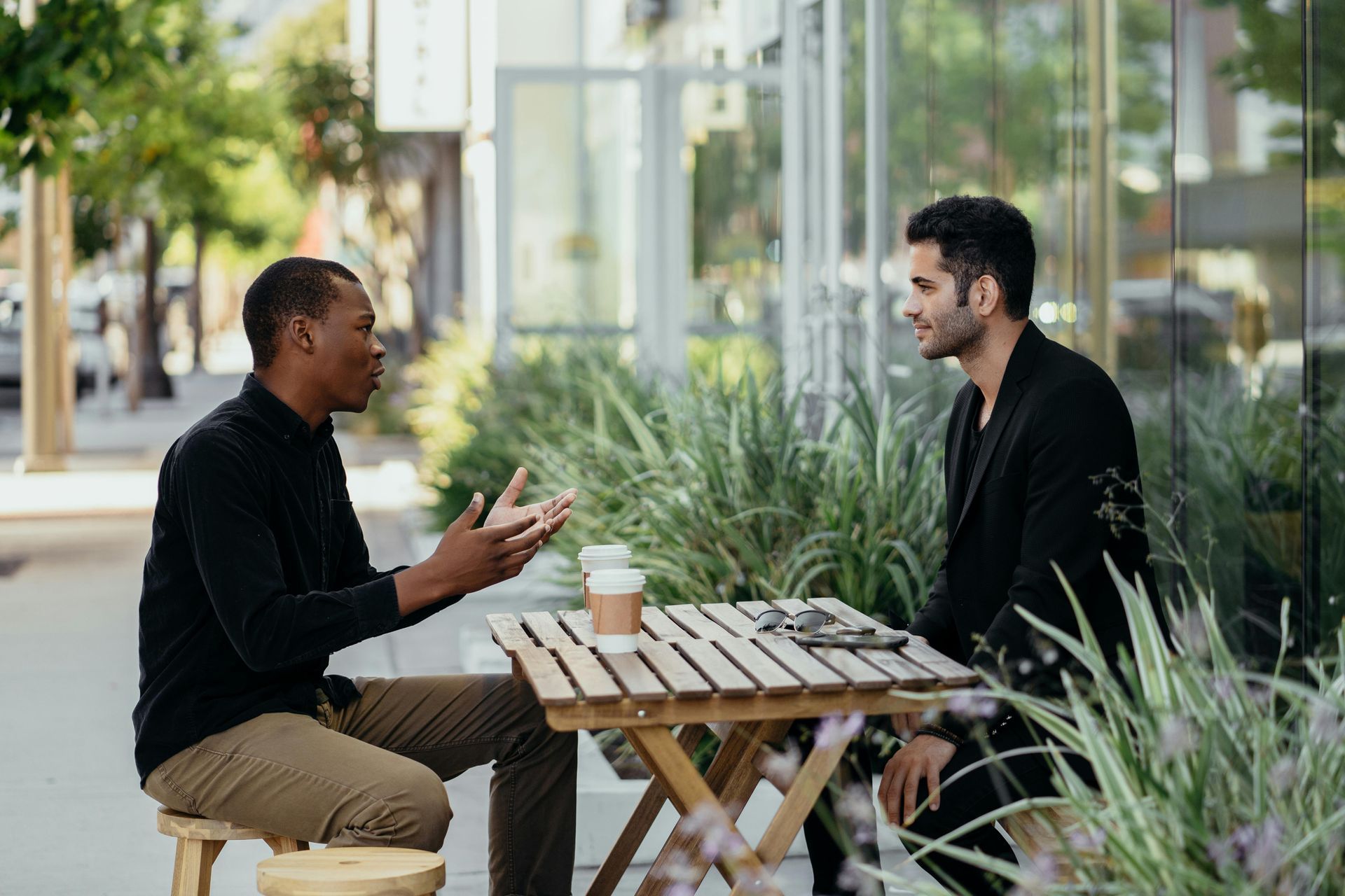 Two men in casual attire seated at a small wooden table outside a building, engaged in conversation. One gestures with his hands while speaking.