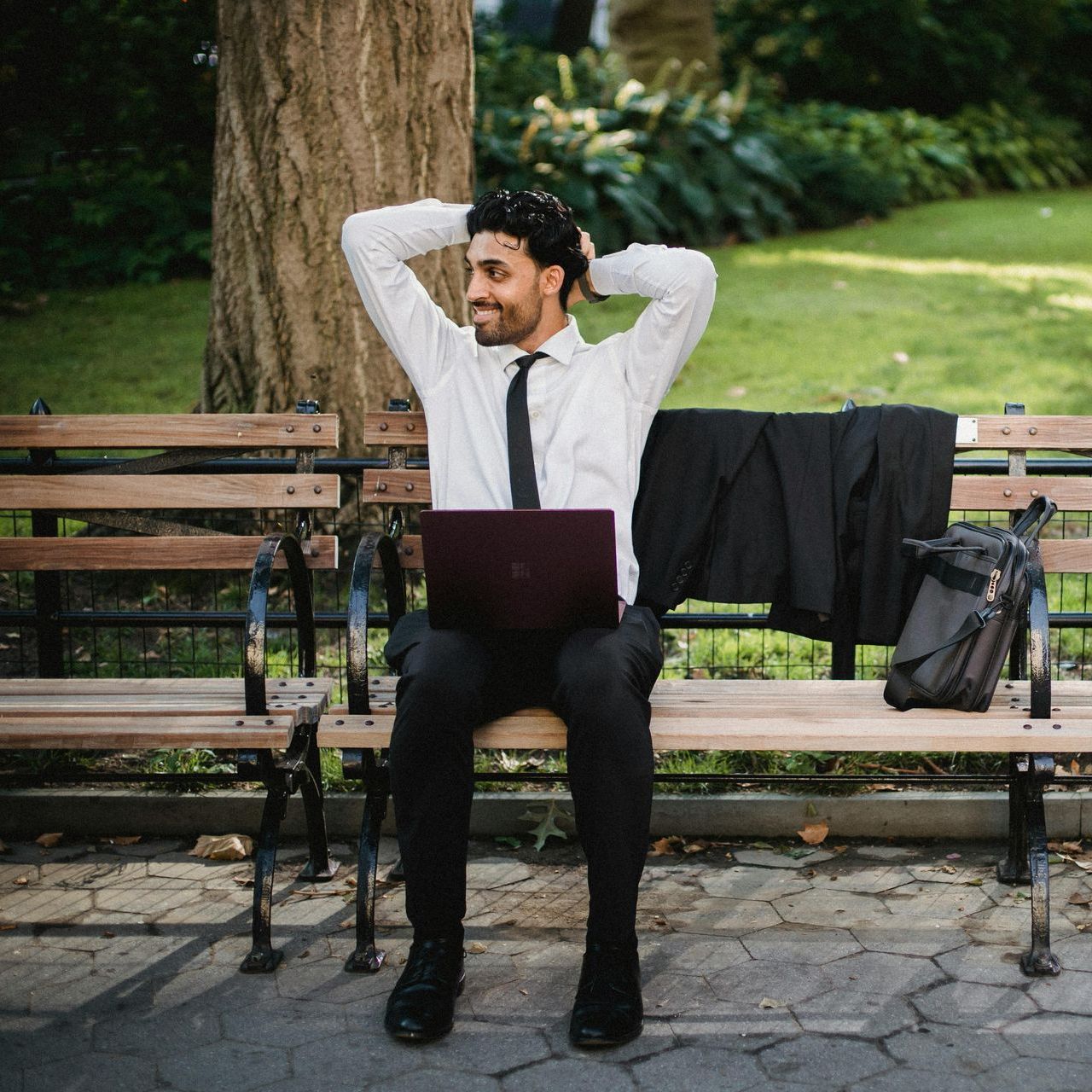 Man in business attire sits on a park bench with a laptop, relaxed, looking to his right. A jacket and bag are next to him.