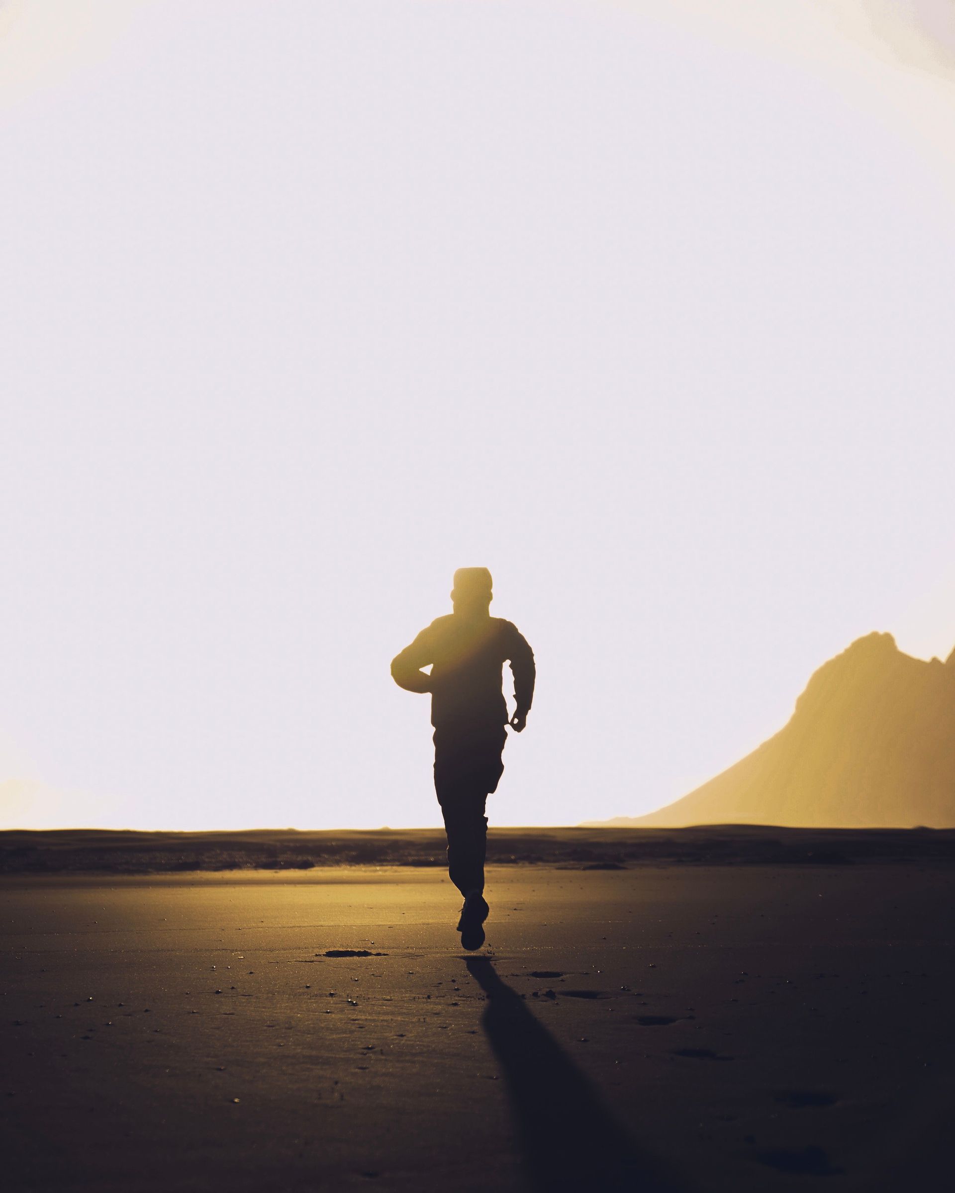 Silhouette of a person running on a beach at sunset, with a rocky hill in the distance.