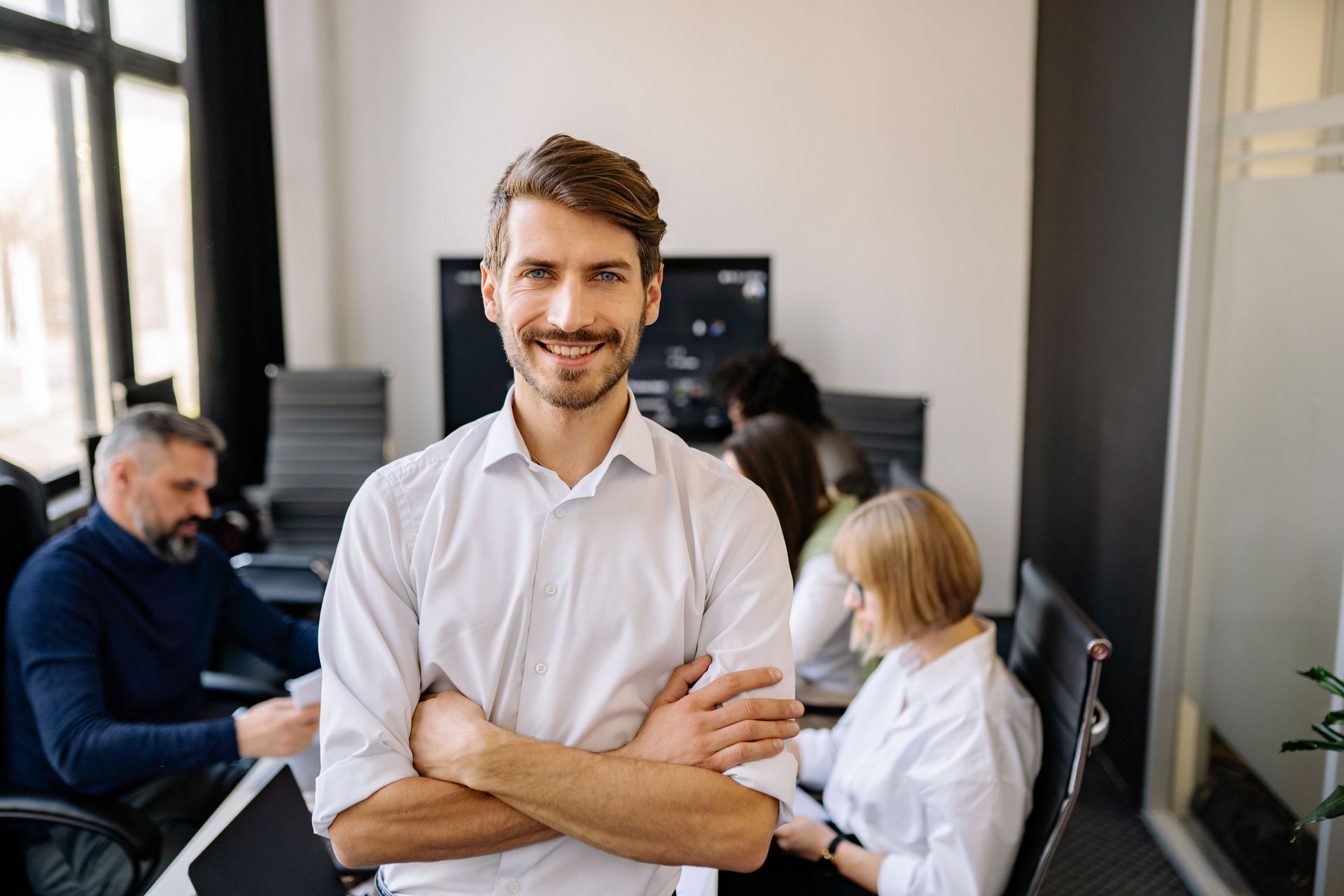 Smiling man in white shirt standing in an office with coworkers in the background