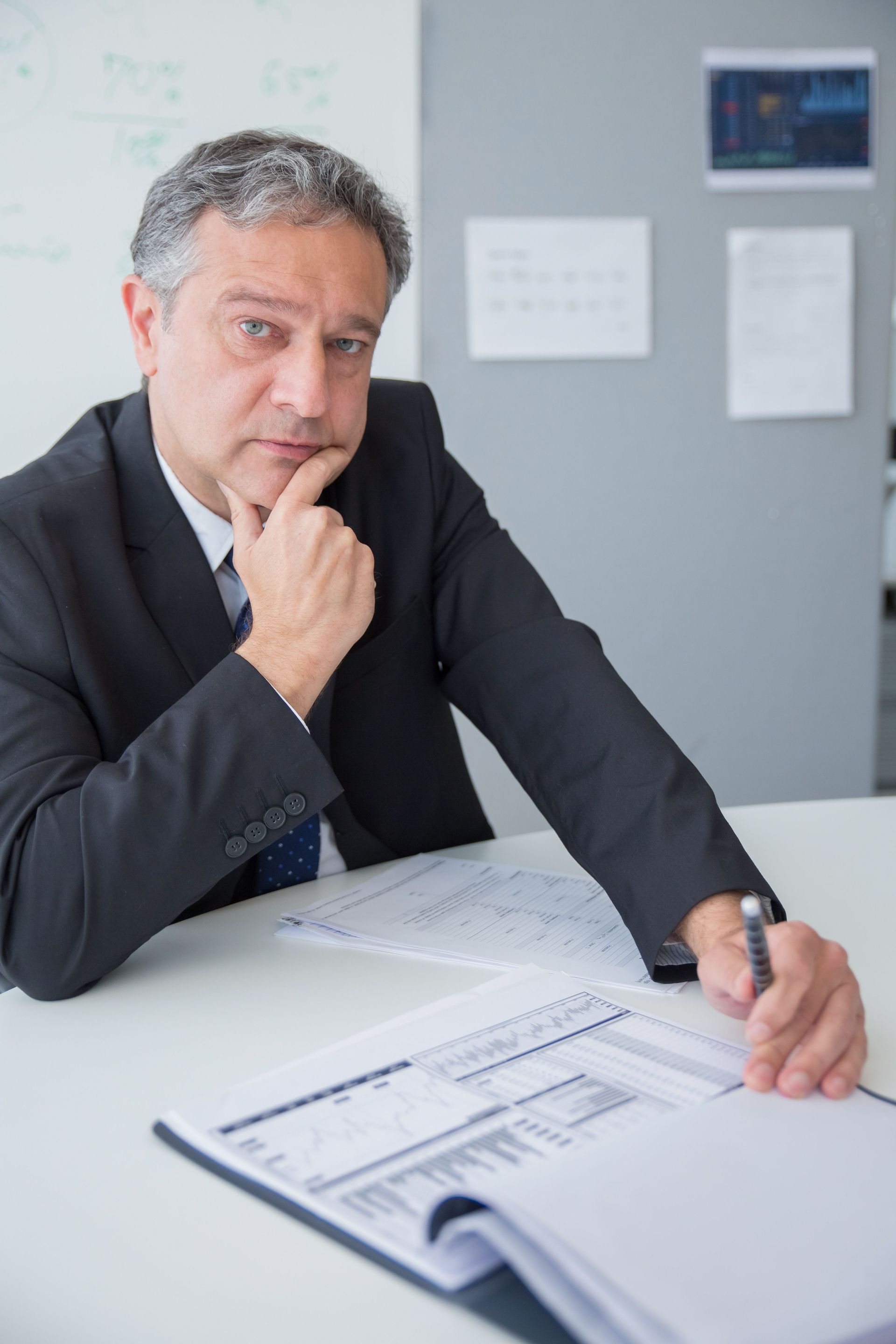 Man in a dark suit leaning over documents at a desk, looking thoughtfully at the camera.