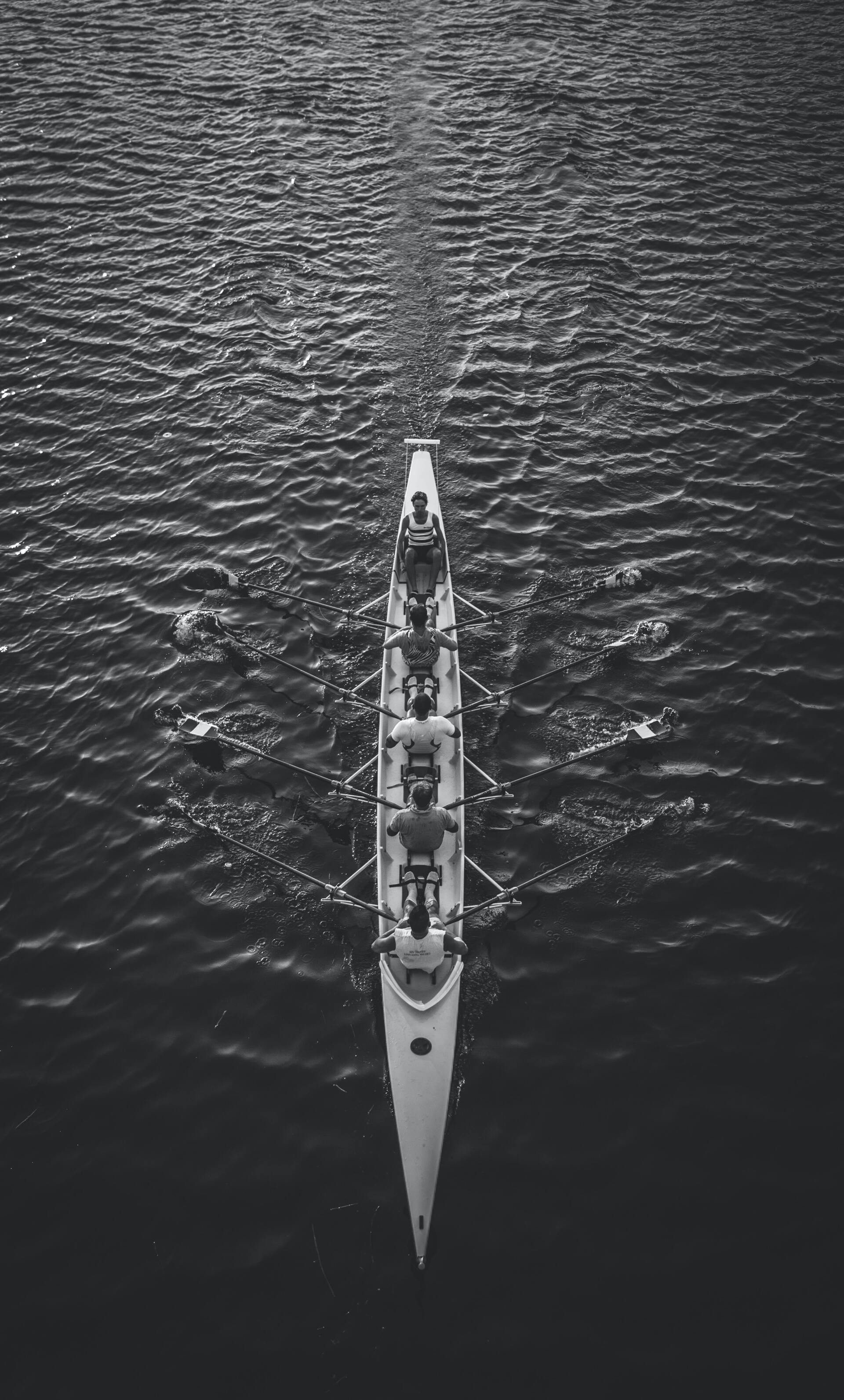 A black and white overhead shot of a rowing team on a lake. Four rowers propel the boat, creating a wake.