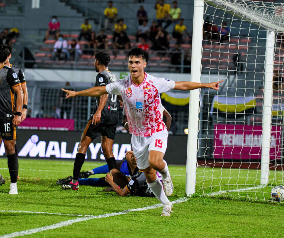 Soccer player in a white jersey celebrates a goal, arms outstretched, near the goalpost on a green field.
