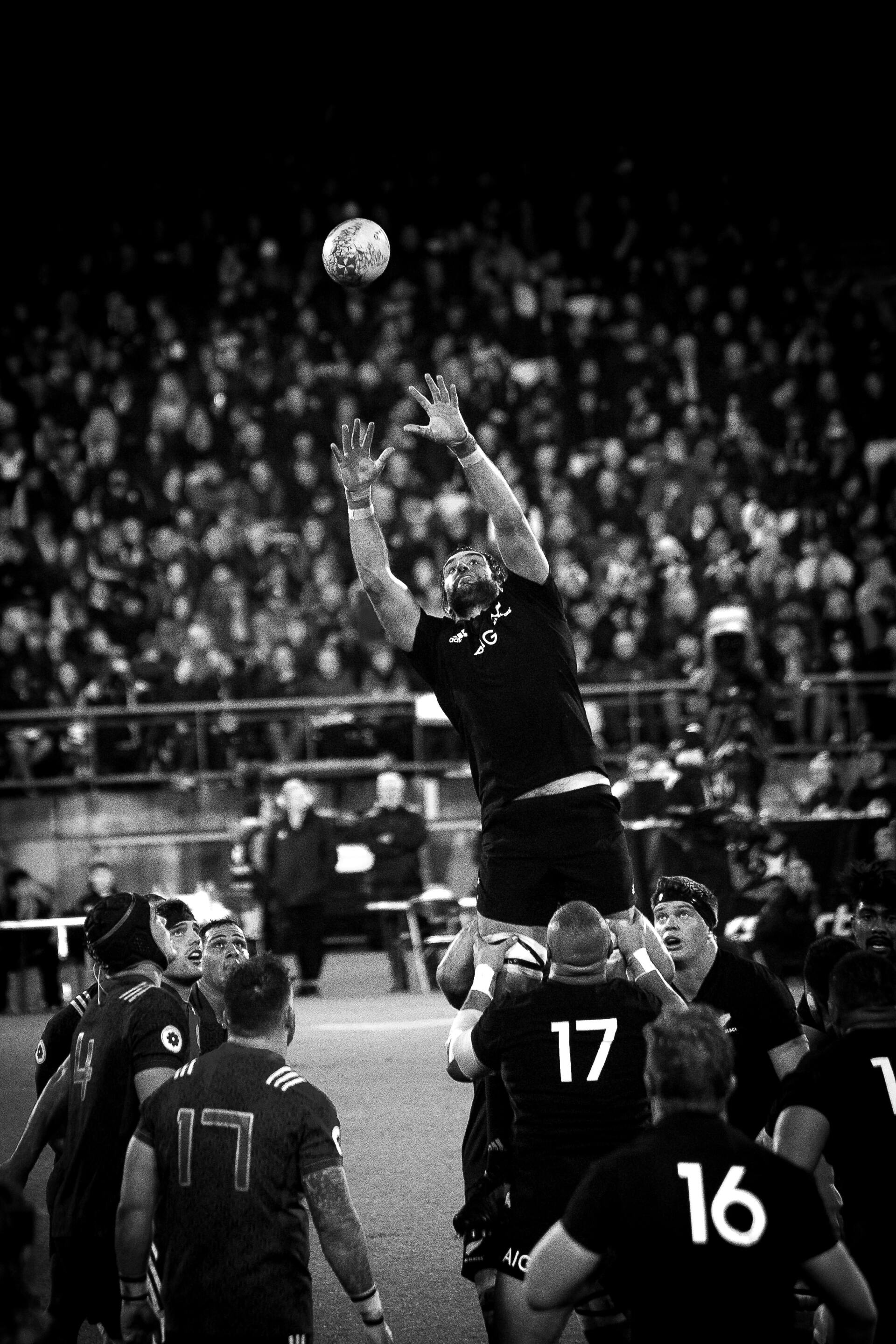Rugby player in black jersey leaping to catch a ball during a game, surrounded by other players and a crowd in a stadium.