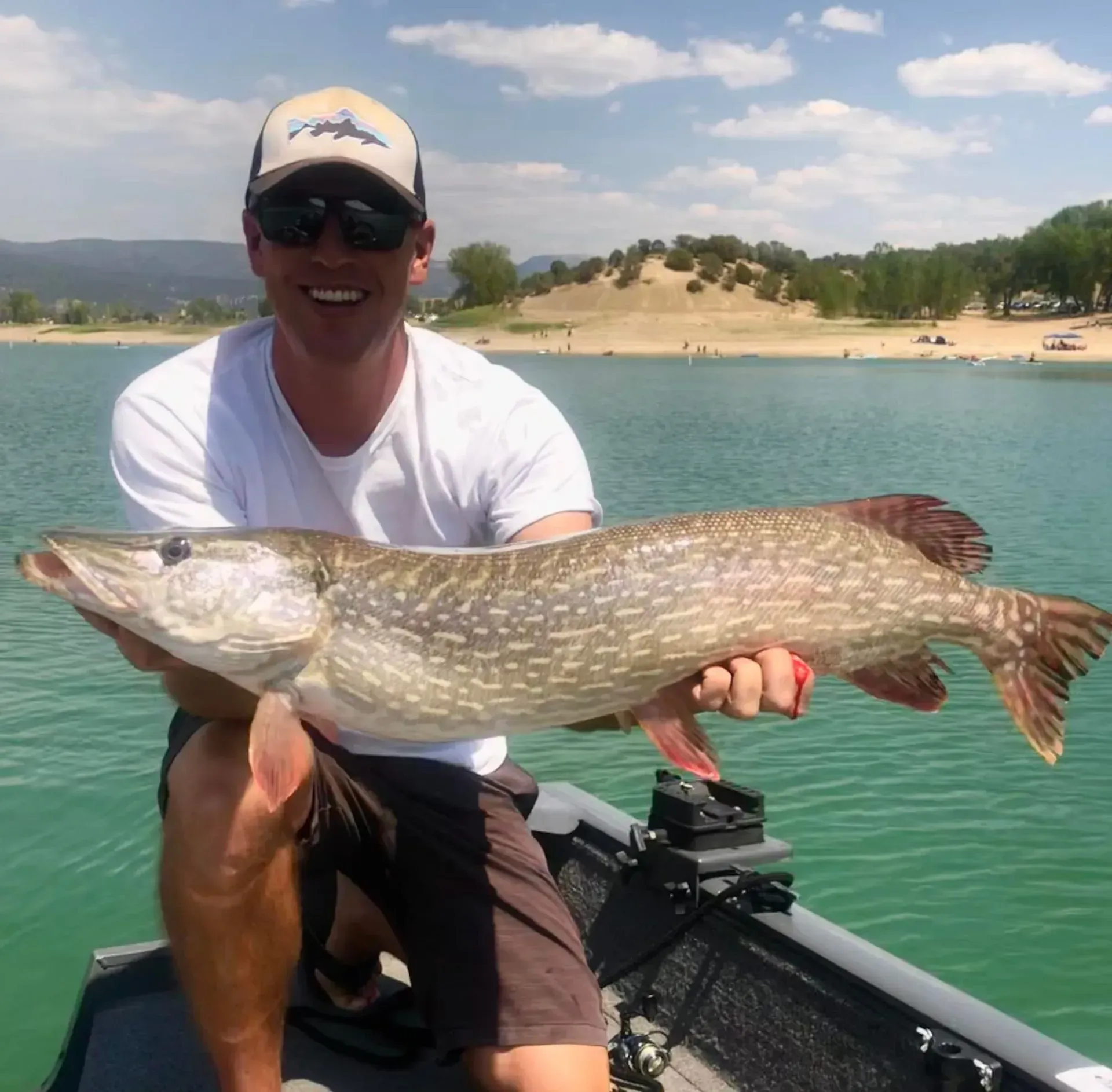 Man in sunglasses holding a large fish, smiling, near a lake.