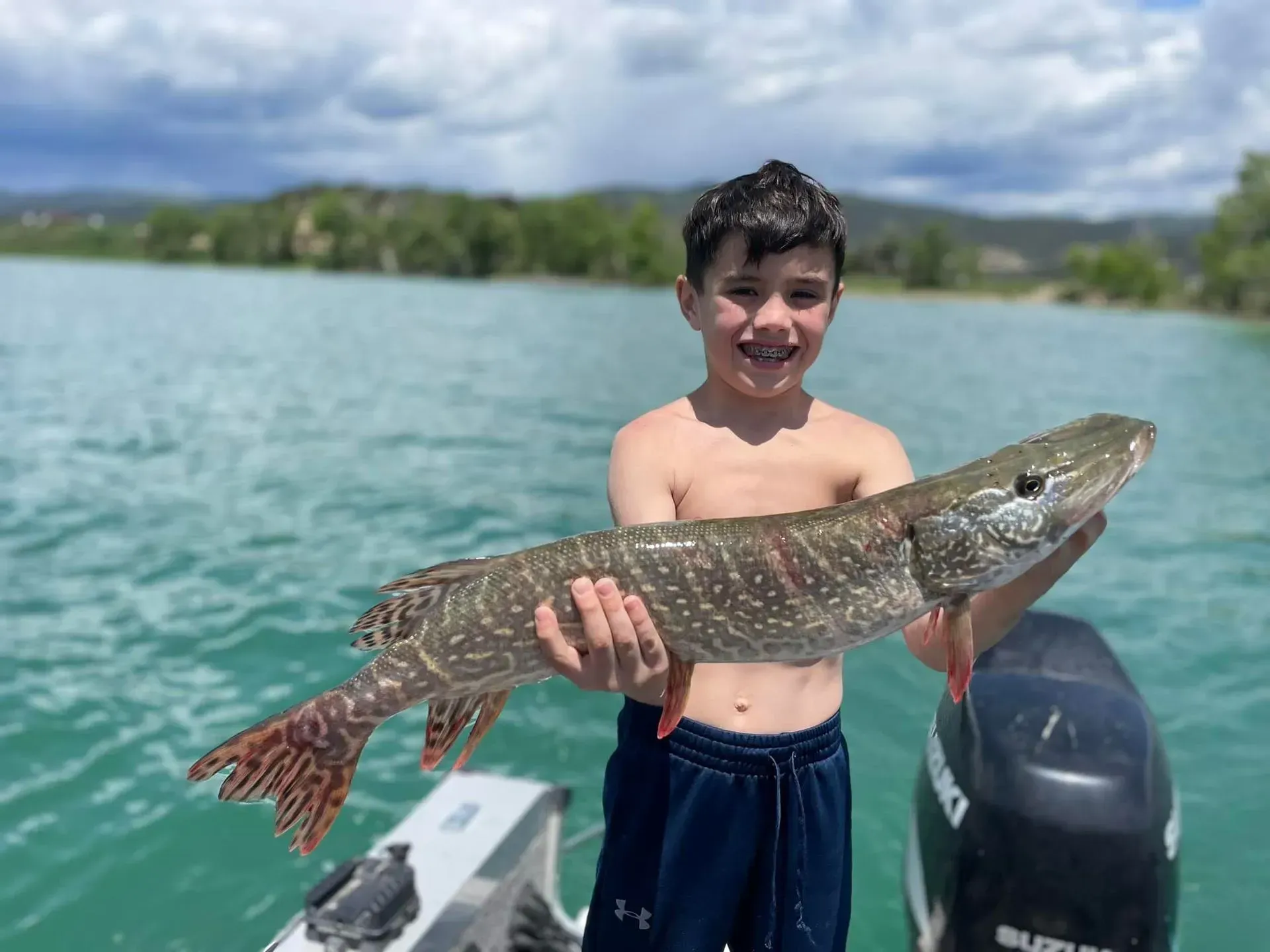 Boy holding a large fish on a boat, turquoise water, trees in the background.