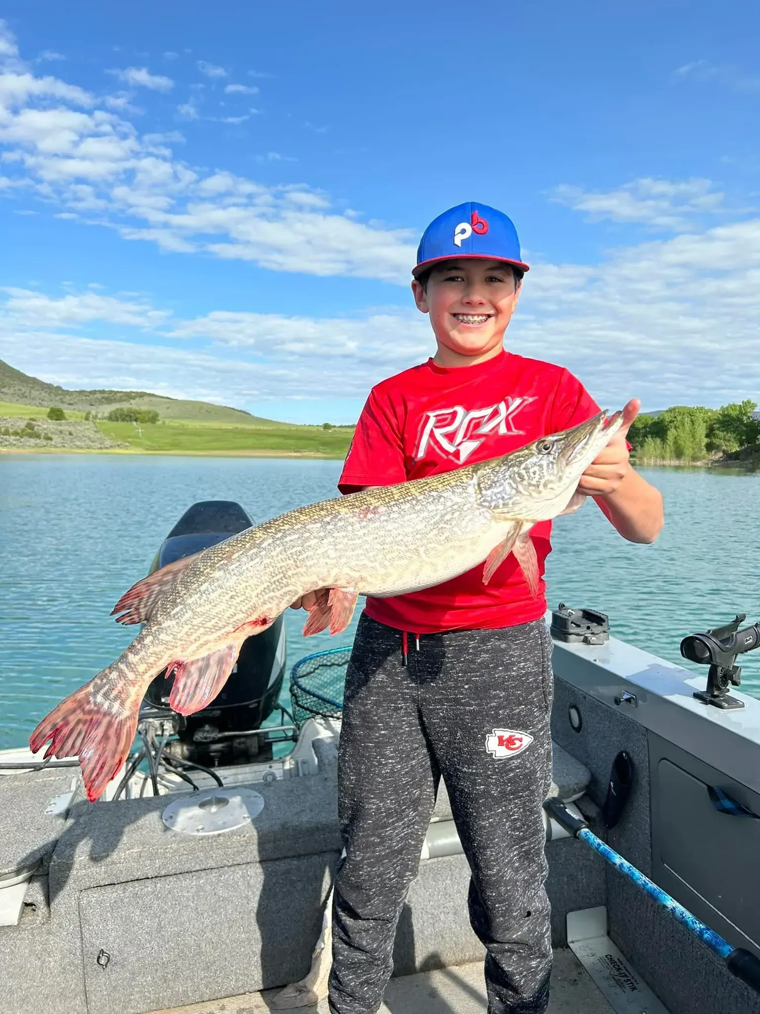 Boy on a boat smiles while holding a large fish, blue sky and lake in the background.