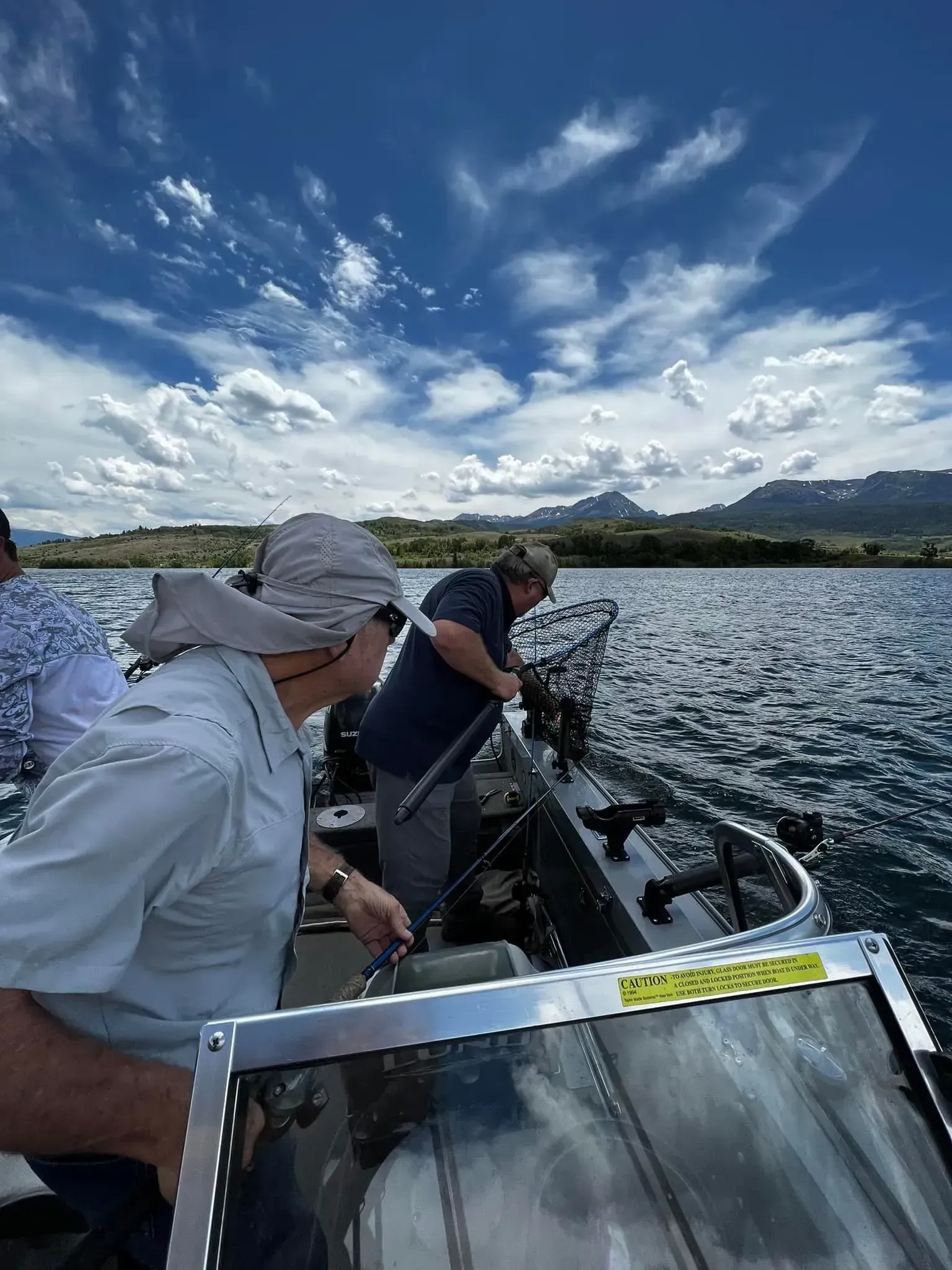 Two people fishing from a boat on a lake under a cloudy blue sky.