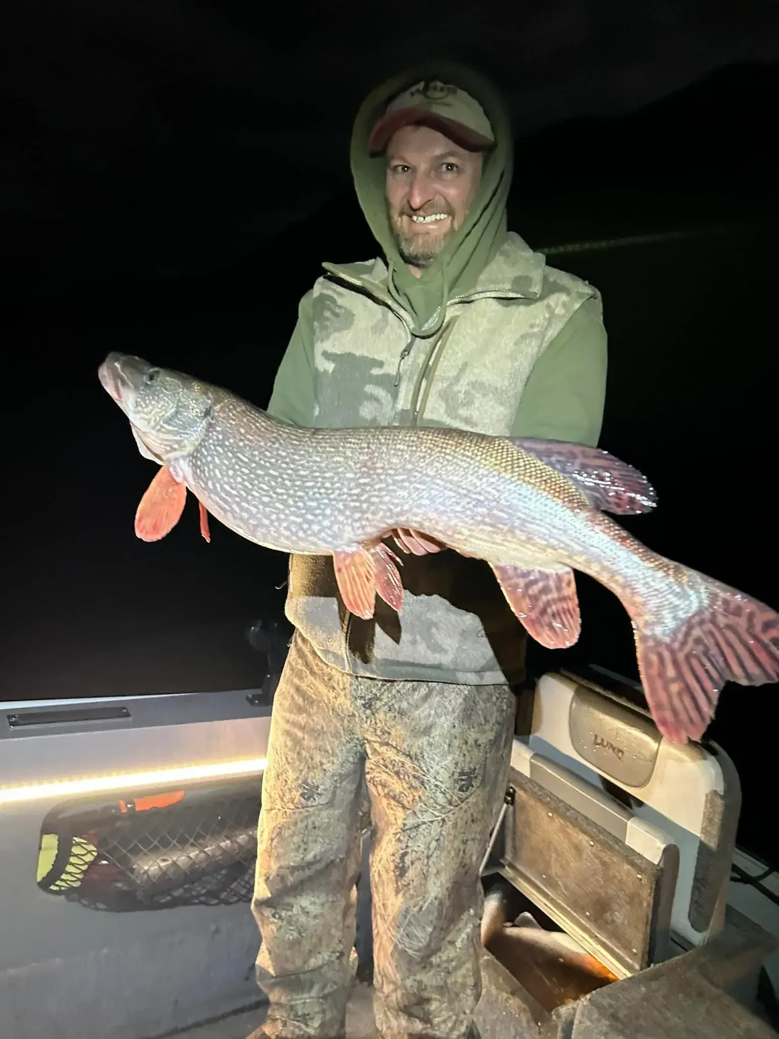 Man in camouflage holding a large fish on a boat at night; smiling.