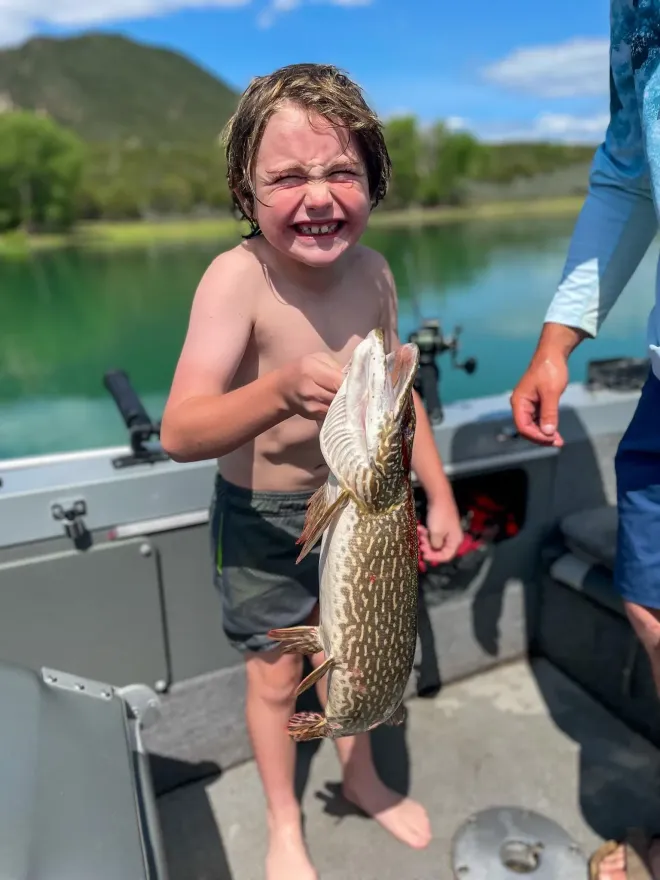 Boy on a boat proudly holding a fish with an open mouth; green water, blue sky background.