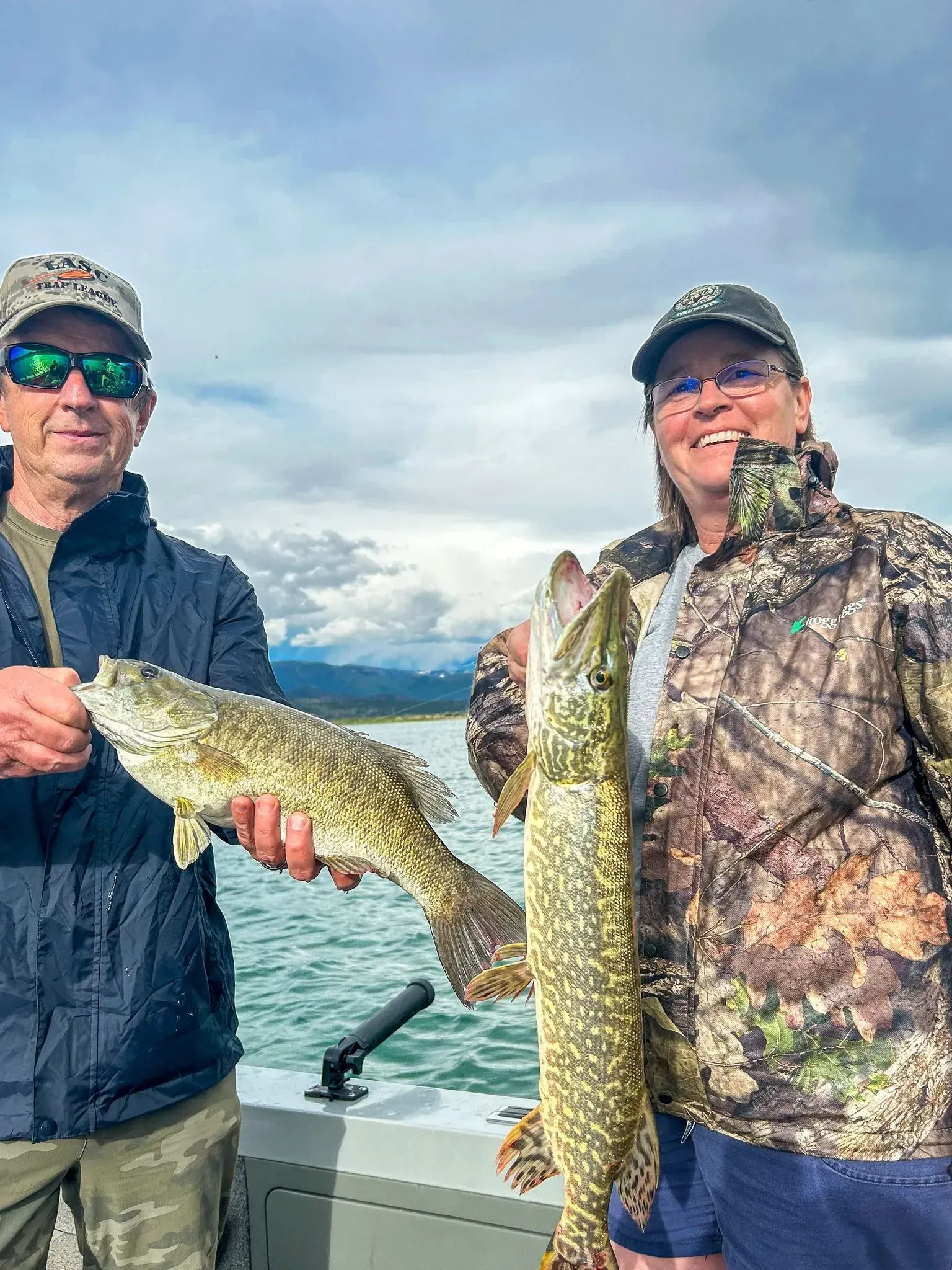 Two people on  on right with a long, green fish, water and mountains in background.